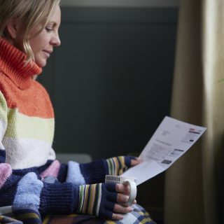 Woman In Gloves With Hot Drink And Bill Trying To Keep Warm By Radiator During Cost Of Living Energy Crisis