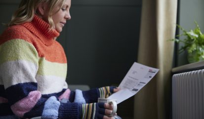 Woman In Gloves With Hot Drink And Bill Trying To Keep Warm By Radiator During Cost Of Living Energy Crisis