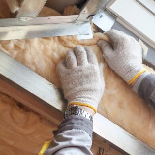 Cropped view on vertical photo with professional worker in protective gloves putting mineral rockwool insulation in wall near windows, behind wooden plank boards. Concept of house under construction